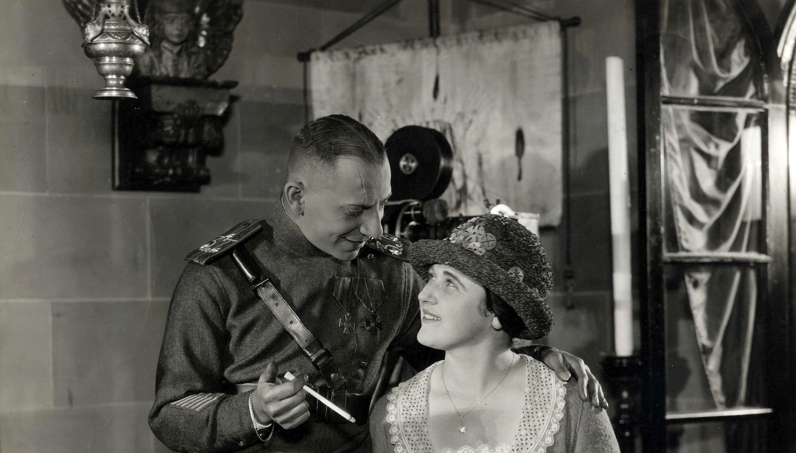 A man in military uniform holding a long cigarette looks down and smiles at a woman in a hat he stands over