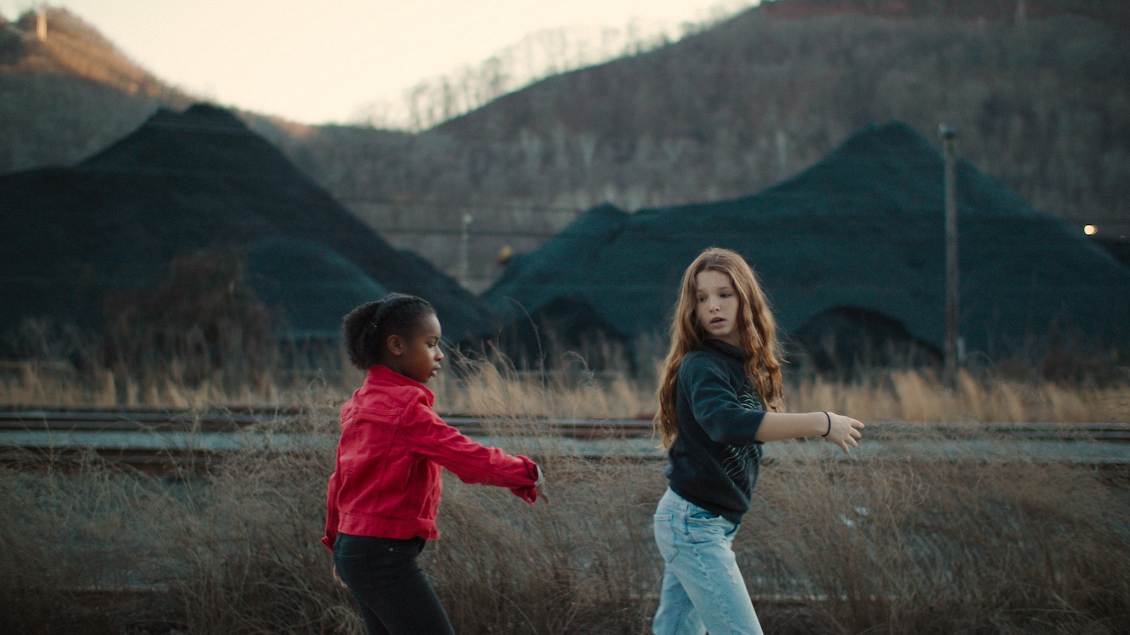 A Black girl in a red jacket and a white girl in a blue jacket together walk against the backdrop of mountains