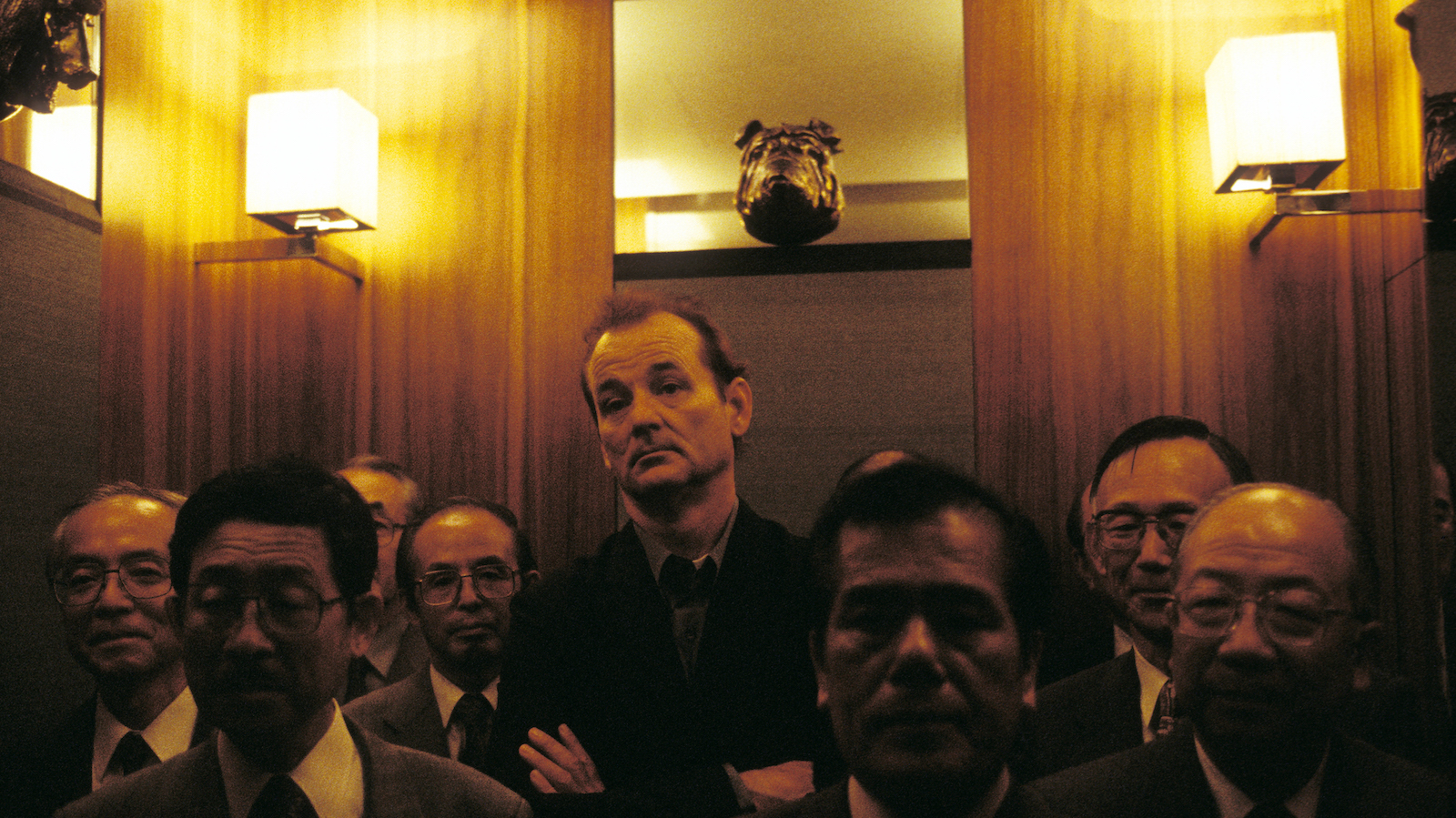 A man stands in an elevator surrounded by men who are shorter than him, his arms folded indifferently