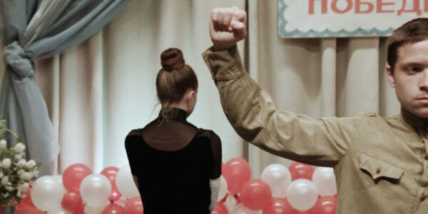 A woman faces away from the camera in a room set for a celebration while a man in a soldier's uniform raises his arm
