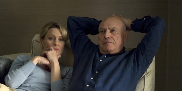 A man and woman lie in bed, both wearing blue shirts. His arms are behind his head and her hand is cradling her chin. Both are pensive.