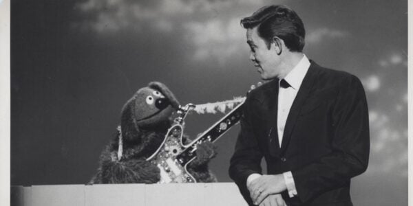 The dog muppet Rowlf holds a guitar and looks up at a man in a suit in a black-and-white image.
