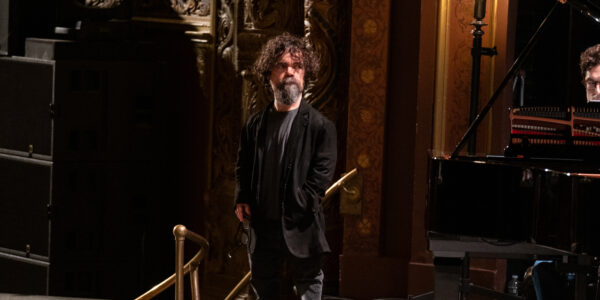 A little person wearing a beard stands backstage at a theater, one hand in his pocket, his hair curly and messy.