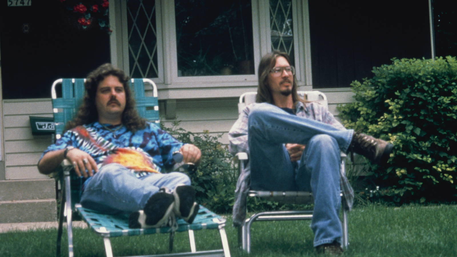 A young man with long hair and glasses and another man with long hair sit on lawn chairs