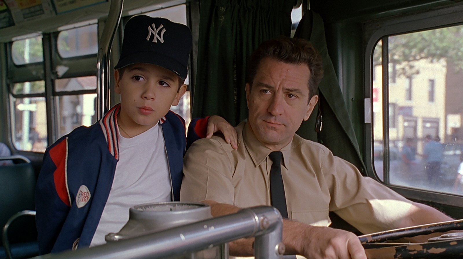 A bus driver in the driver's seat sits next to a young boy in a New York Yankees cap, who stands with his hand on the man's shoulder
