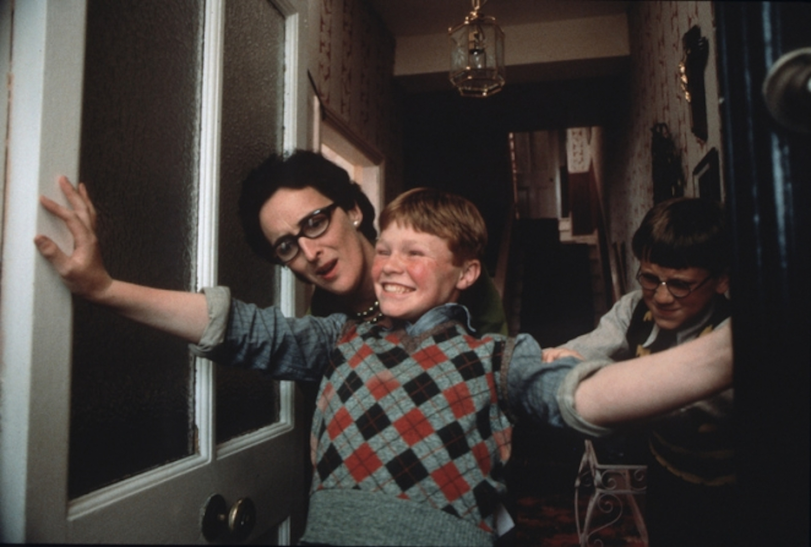 A red-headed boy in an argyle vest smilingly holds his arms across a doorway as two others peek out behind him