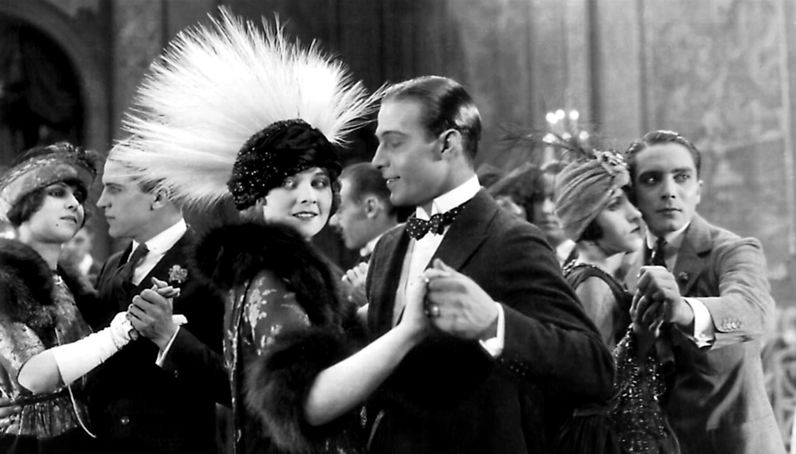 A black and white image of a man in a tuxedo dancing with a woman in a high white feathered peacock headdress