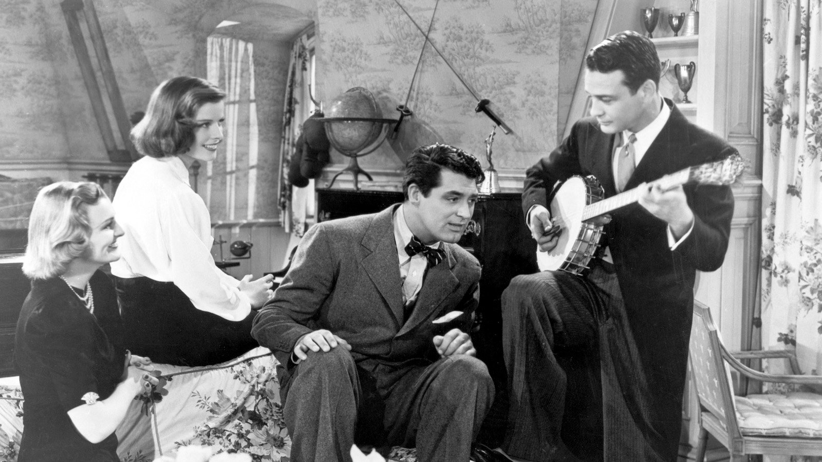A man standing and playing a banjo on the right of the frame, with another man perched on a couch leaning his head in to listen to him and two women sitting on the other side of the couch