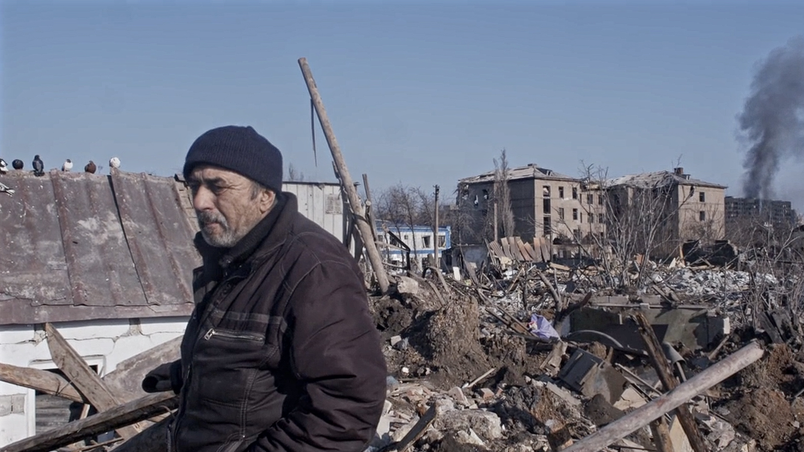A serious looking man in a stocking cap walks before building rubble