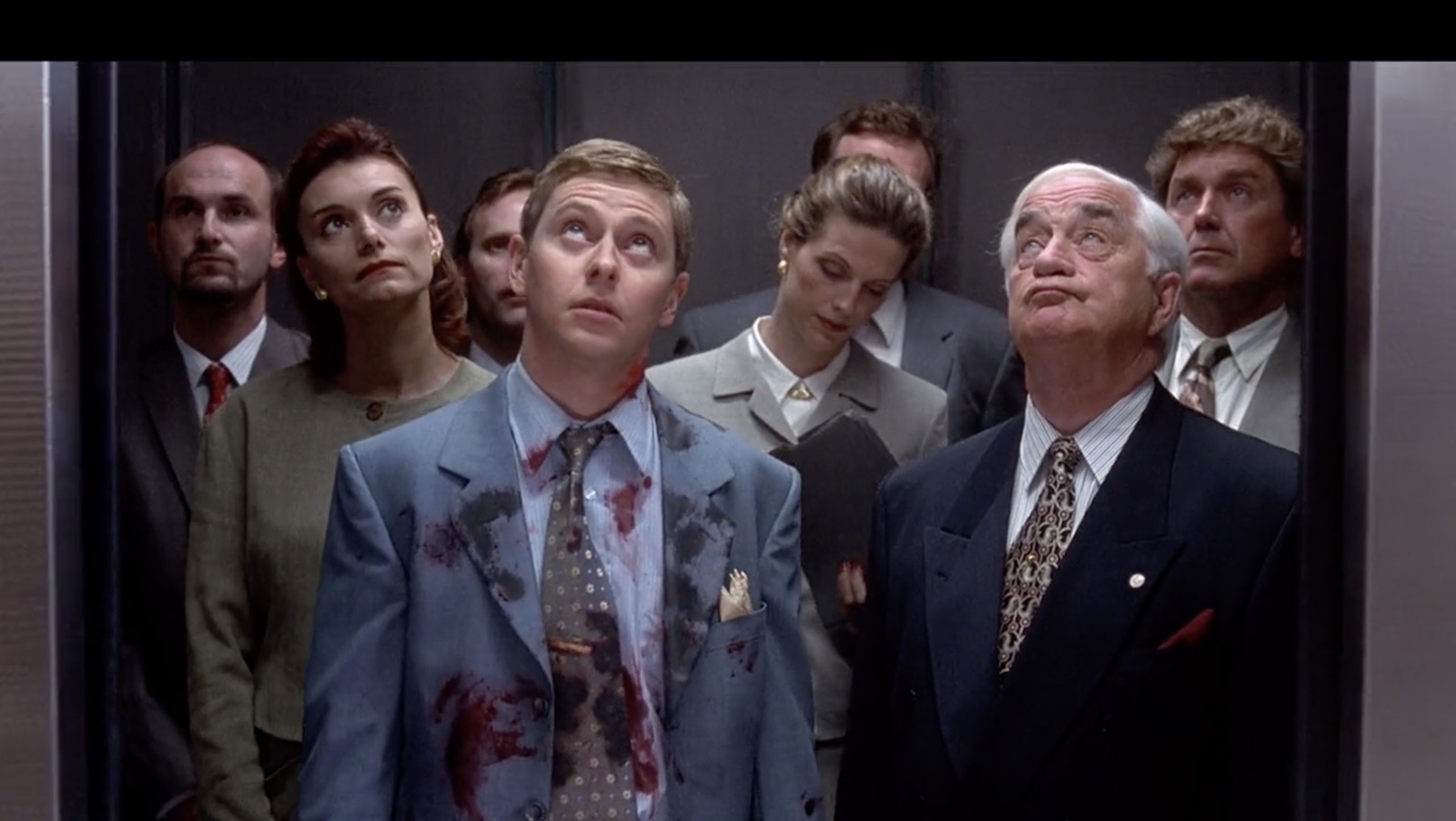 A man in a bloodied suit stands in an elevator and looks up, surrounded by others