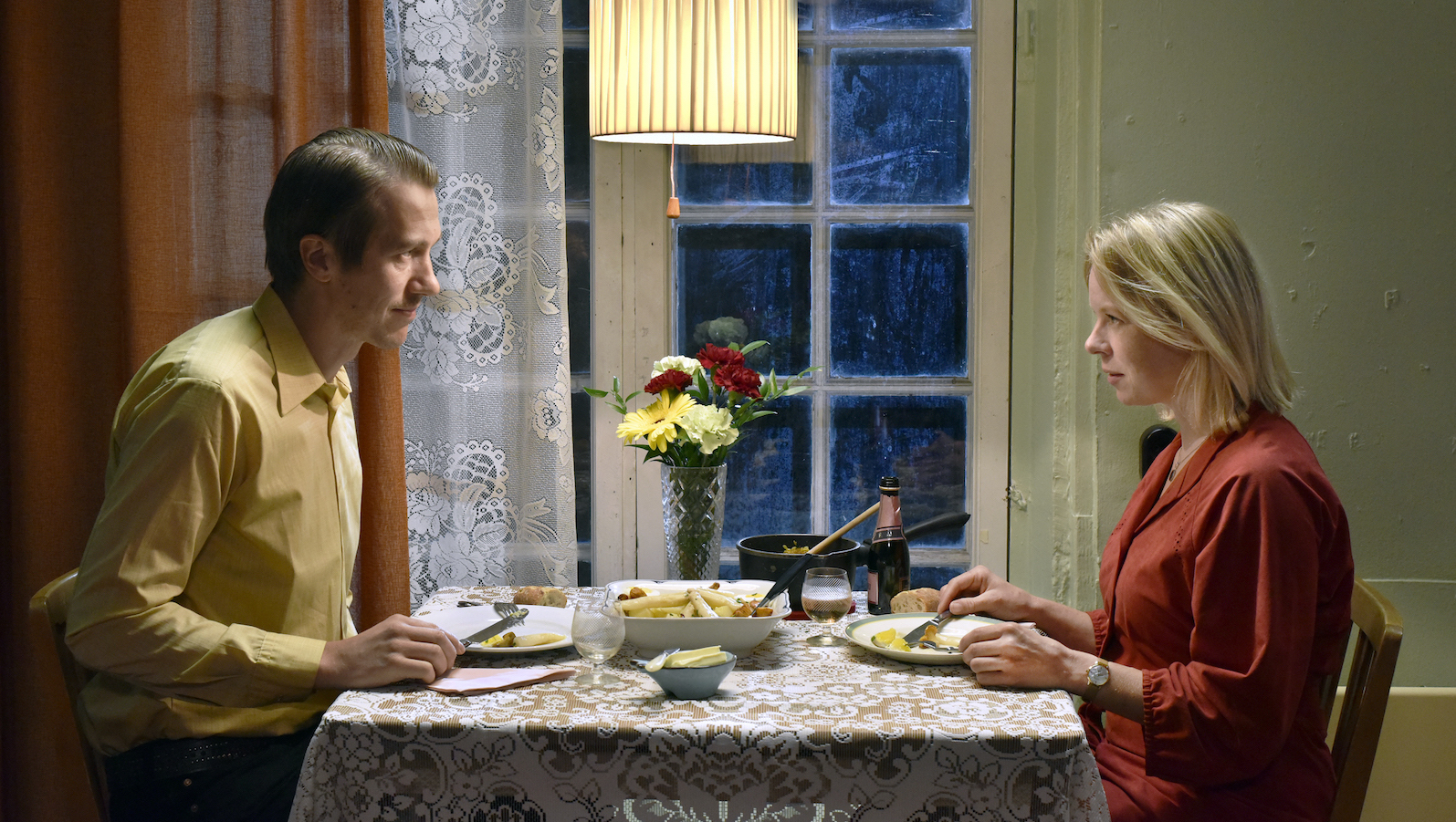 A man and woman sit at a table with flowers on it having dinner and facing each other