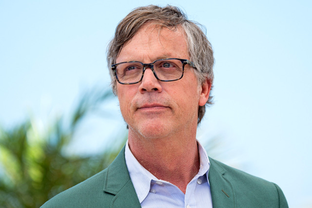 A white man wearing glasses, a blue shirt and green jacket, seen in a head shot against a blue sky