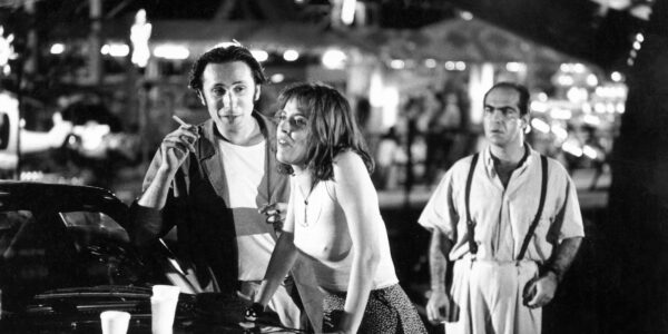 Three people stand by a car in an outdoor carnival, a woman leaning against the car hood and a man holding a cigarette talking to her, while another man stands behind them and looks at them
