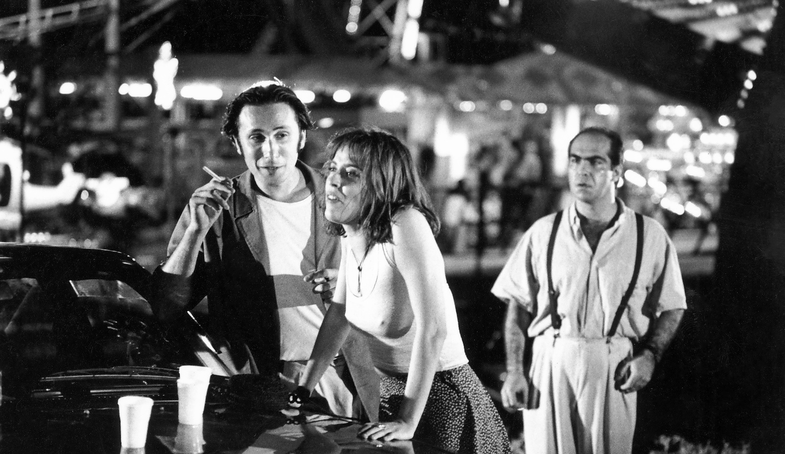 Three people stand by a car in an outdoor carnival, a woman leaning against the car hood and a man holding a cigarette talking to her, while another man stands behind them and looks at them