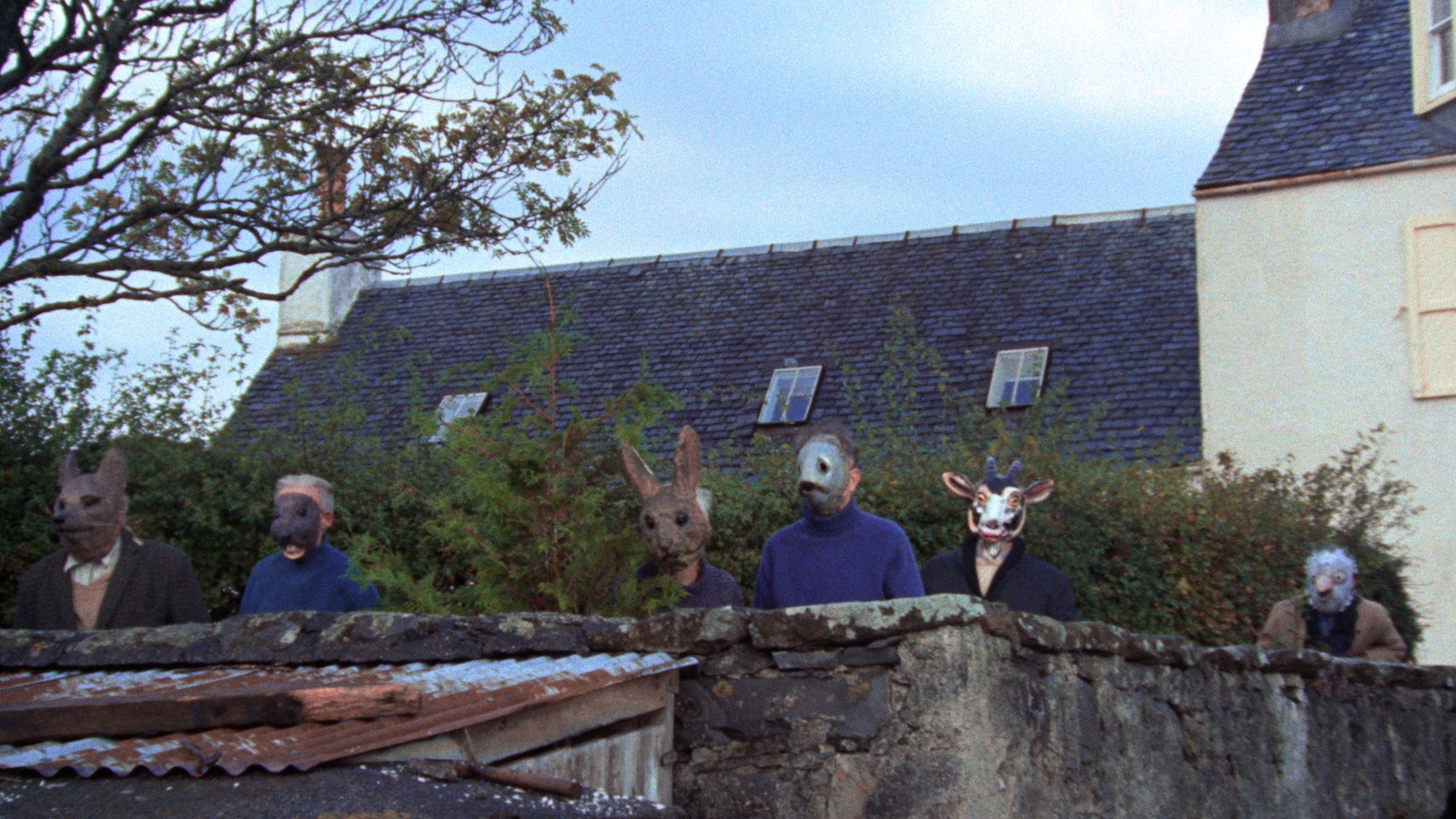 People in animal masks stand behind a long stone wall behind a rural house