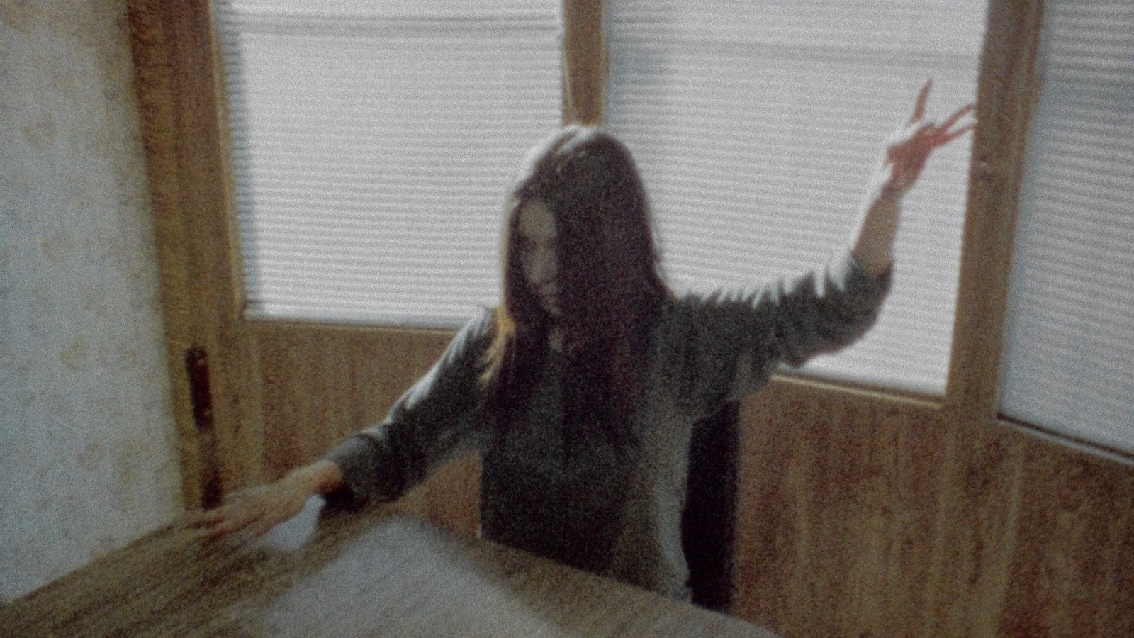 A woman with long hair sits at a table and raises one hand in the air and the other hovers above the table spread out, her fingers making strange signals