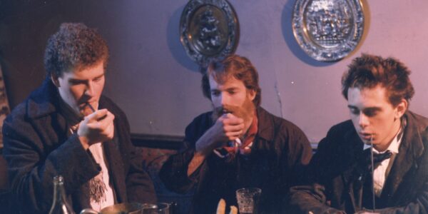 Three men in old-fashioned clothes sit at a table smoking pipes