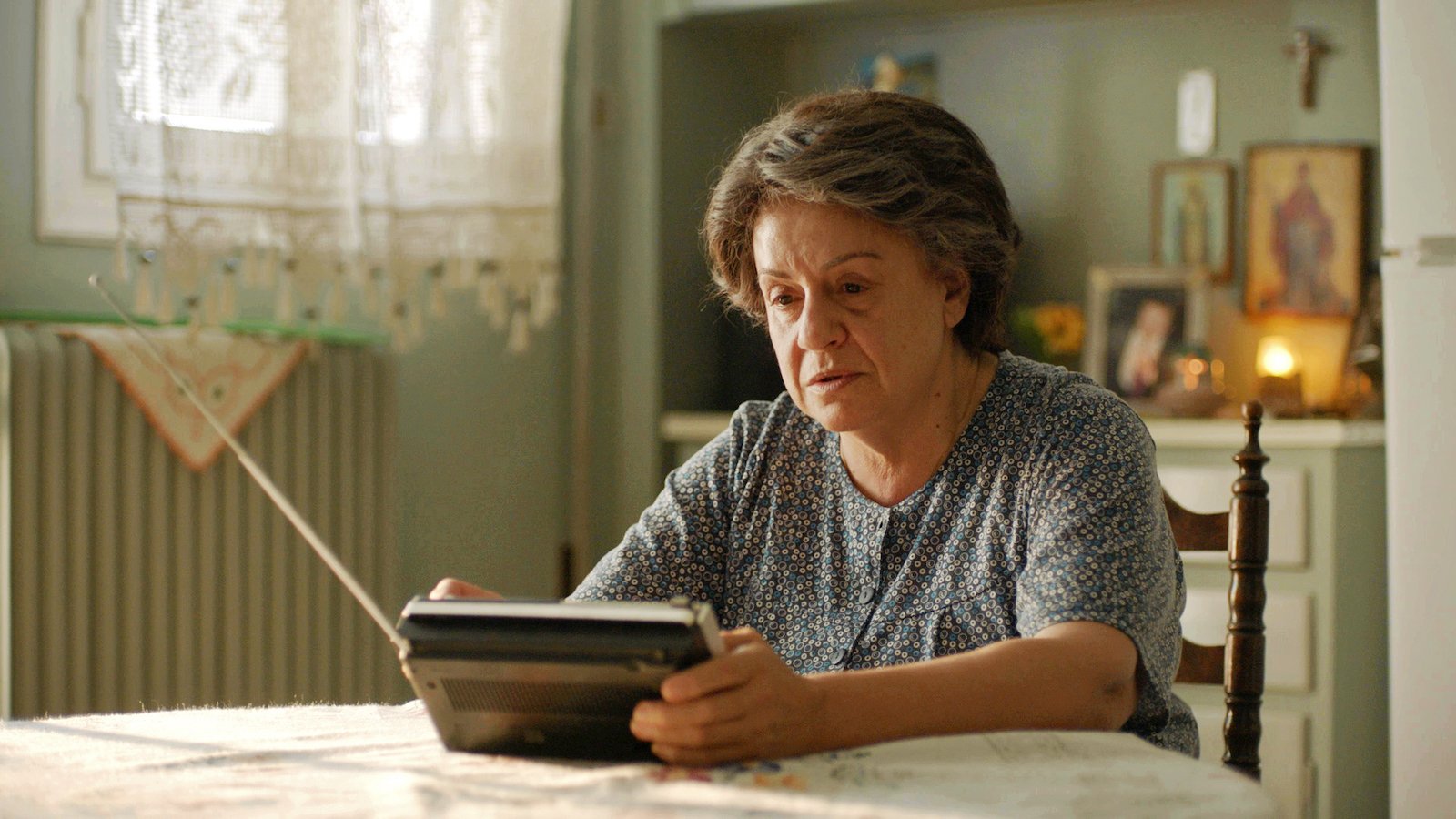 An older woman sits at a kitchen holding a transistor radio