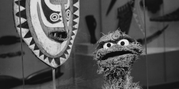 A furry muppet (Oscar the Grouch) looks confused at camera in front of an African mask at a museum