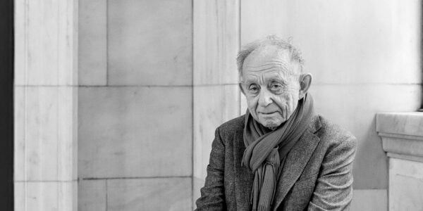 a black and white image of a man looking at the camera wearing a scarf, sitting outside a library wall