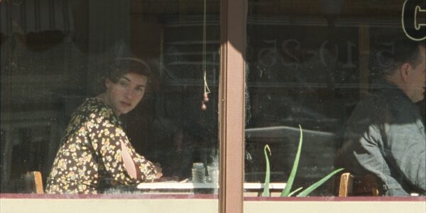 A woman in 1930s dress sits in the window of a diner looking out