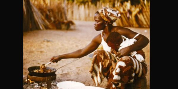 A woman holding a baby in one hand sits before an open fire cooking chicken in a rural African village