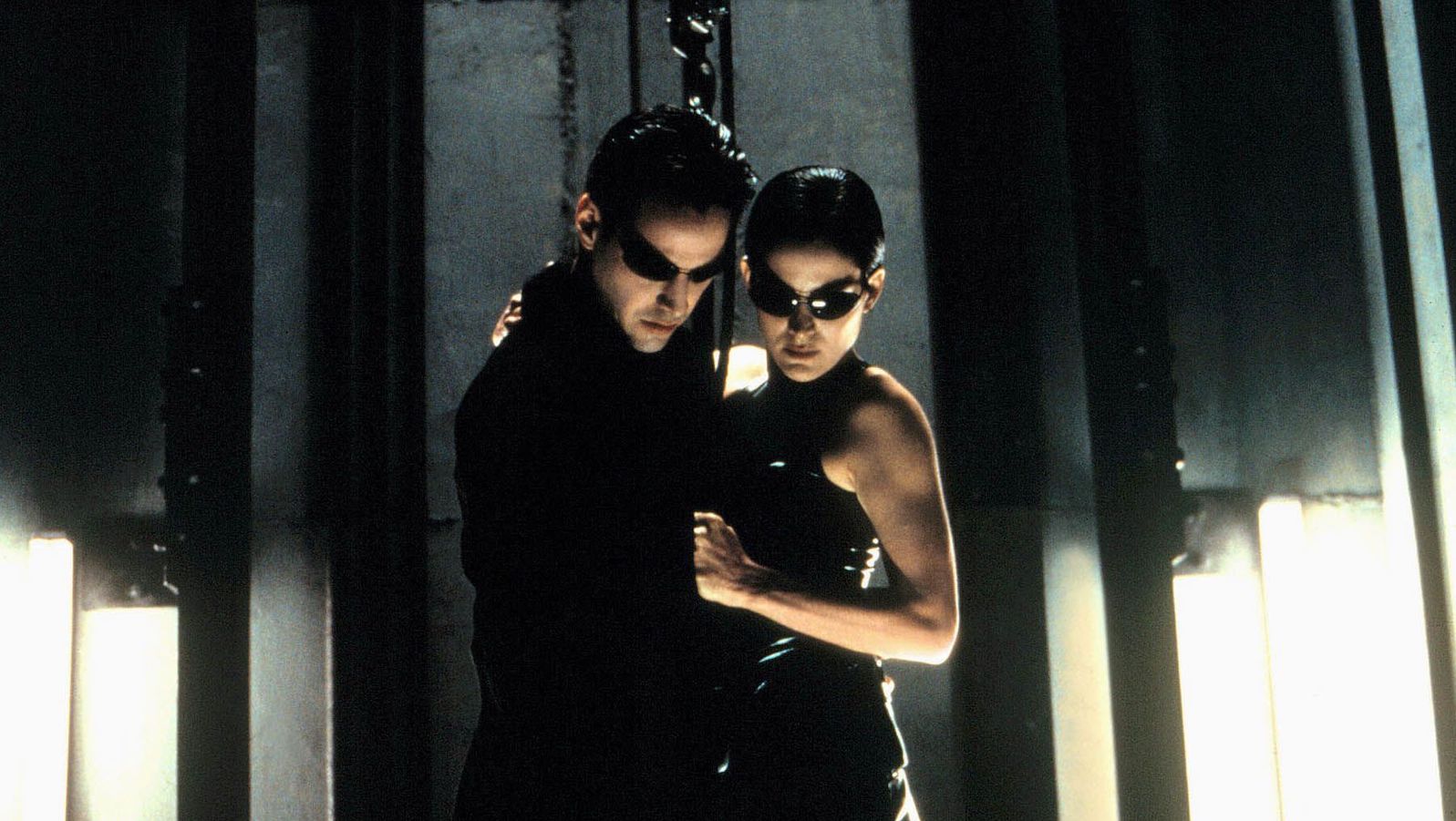 A man and woman in sunglasses and wearing leather hold each other and look down.