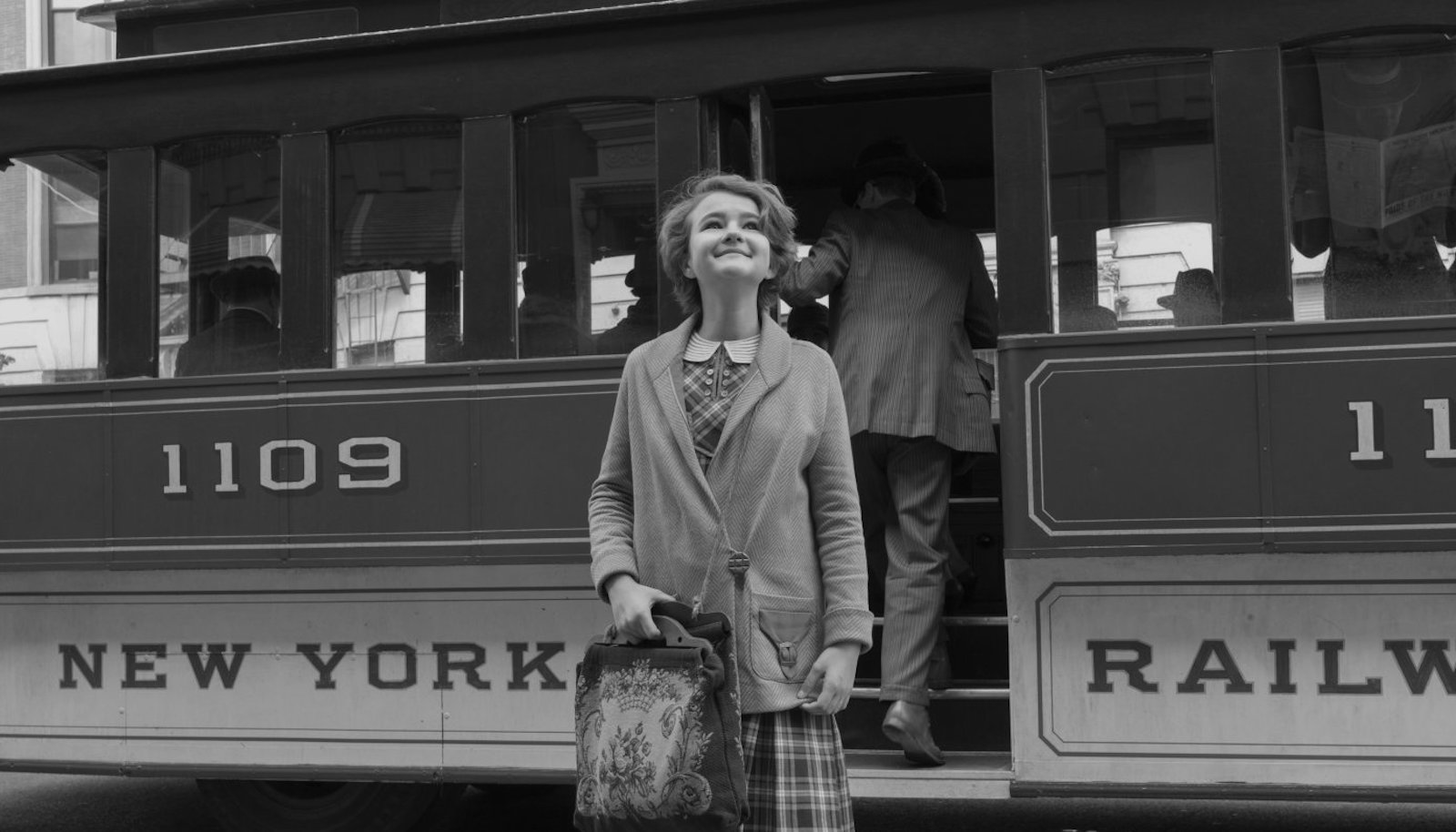 A young girl holding a suitcase has stepped off a train and looks up with hope and excitement in a black and white photo