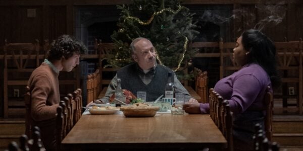 Three people sit around a dinner table talking to each other and eating with a Christmas tree behind the man who sits at the head of the table