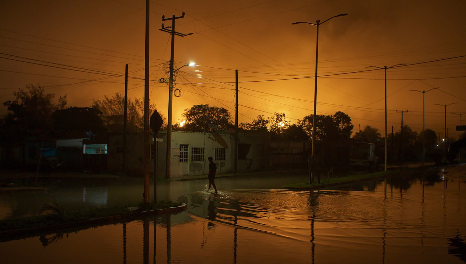 A yellow sky blazes behind a flooded street at night, with a solitary figure walking through water in front of a house.