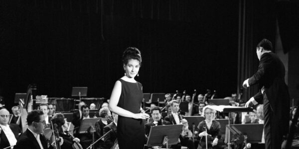 A regal singer in a black dress stands in a symphony space. She is surrounded by an orchestra.