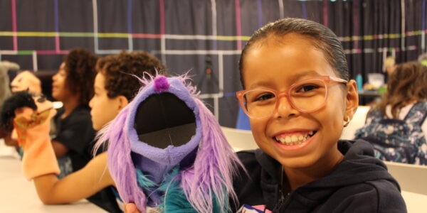 A little girl in glasses holds up a puppet and smiles at camera