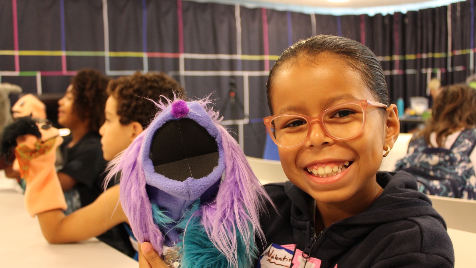 A little girl in glasses holds up a puppet and smiles at camera