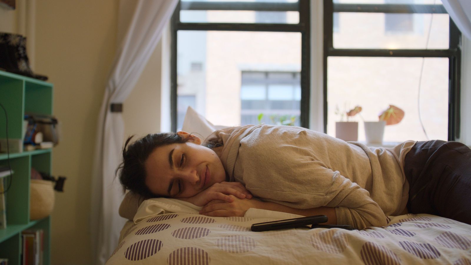A woman lies on a bed during the daytime with her eyes closed and smiling