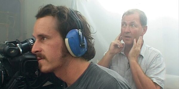 A man holding his fingers in his ears stands behind a man wearing blue sound-canceling headphones