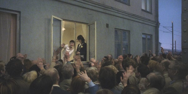 A newly married couple, she in a wedding dress and he in a tux and holding a guitar, stand near an open window in a city apartment waving at a crowd of people on the street