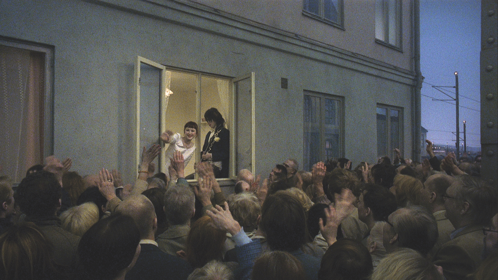A newly married couple, she in a wedding dress and he in a tux and holding a guitar, stand near an open window in a city apartment waving at a crowd of people on the street