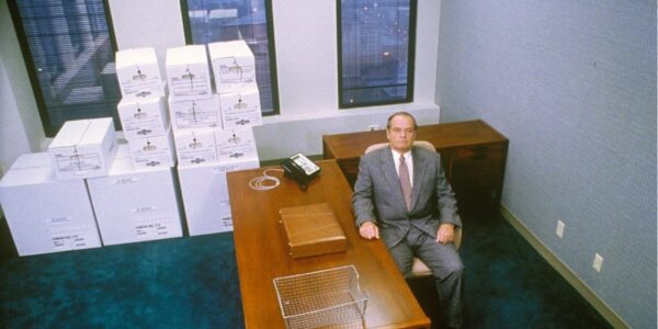 A man sits in a near empty, blank office looking up to the ceiling; his desk is bare except for a telephone, briefcase, and empty metal tray. Behind him are file boxes piled neatly.
