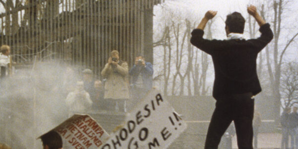 A man stands at a street protest with his arms in the air while people in the distance take photos of him; a sign near him says RHODESIA GO HOME!