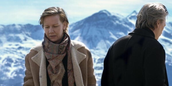 A man and woman stand with a snowy mountain backdrop, she faces forward hm backward