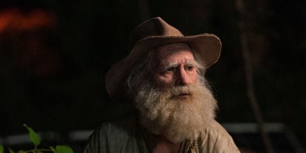 An elderly man with a long white beard and wearing a floppy hat looks off-screen