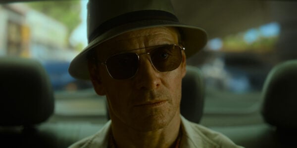 A man in sunglasses and a fedora sits in the back of a car looking straight ahead with a serious expression
