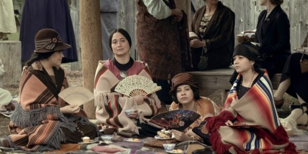 A group of native American women sit on a blanket on the ground, the second from the left holding a hand fan, looking off with a sly interested expression