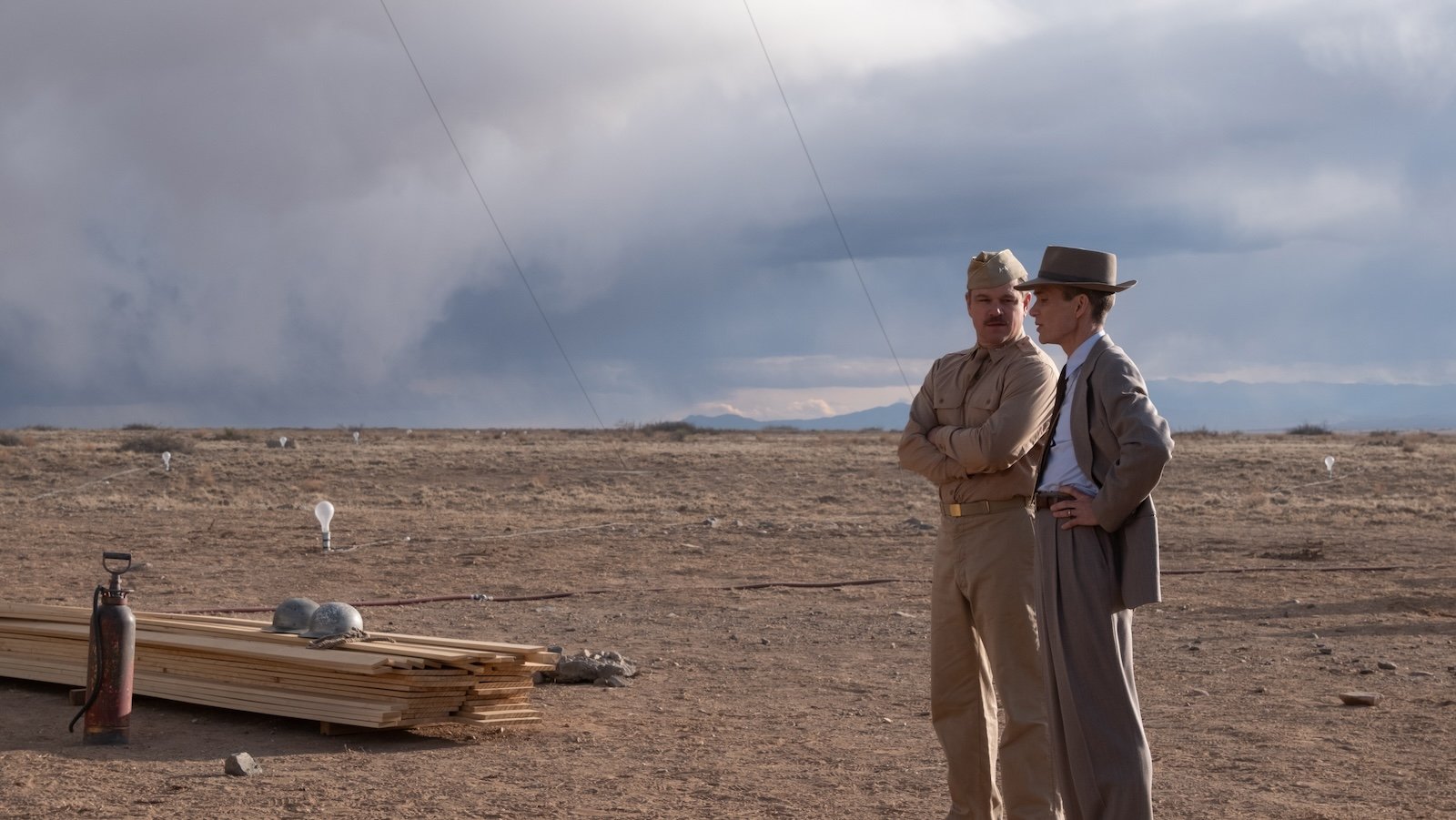 A man in a suit and fedora stands next to a man in military uniform against a vast desert background
