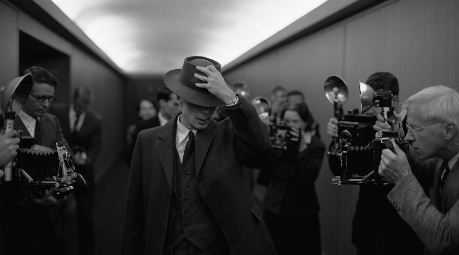 A man covers his face with his fedora as he walks down a hallway of reporters taking photographs in a black and white image