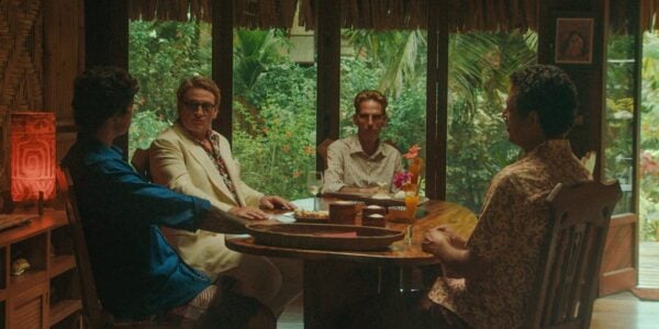 Four men sit around a table in tropical-colored clothes with dense green jungle leaves outside the window