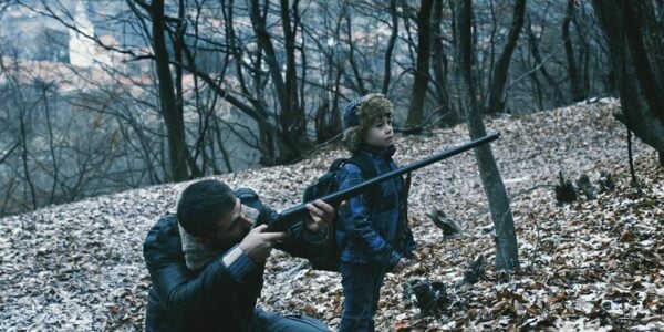 A man and a boy in the woods, surrounded by bare trees and leaves on the ground; the man aims a rifle off screen