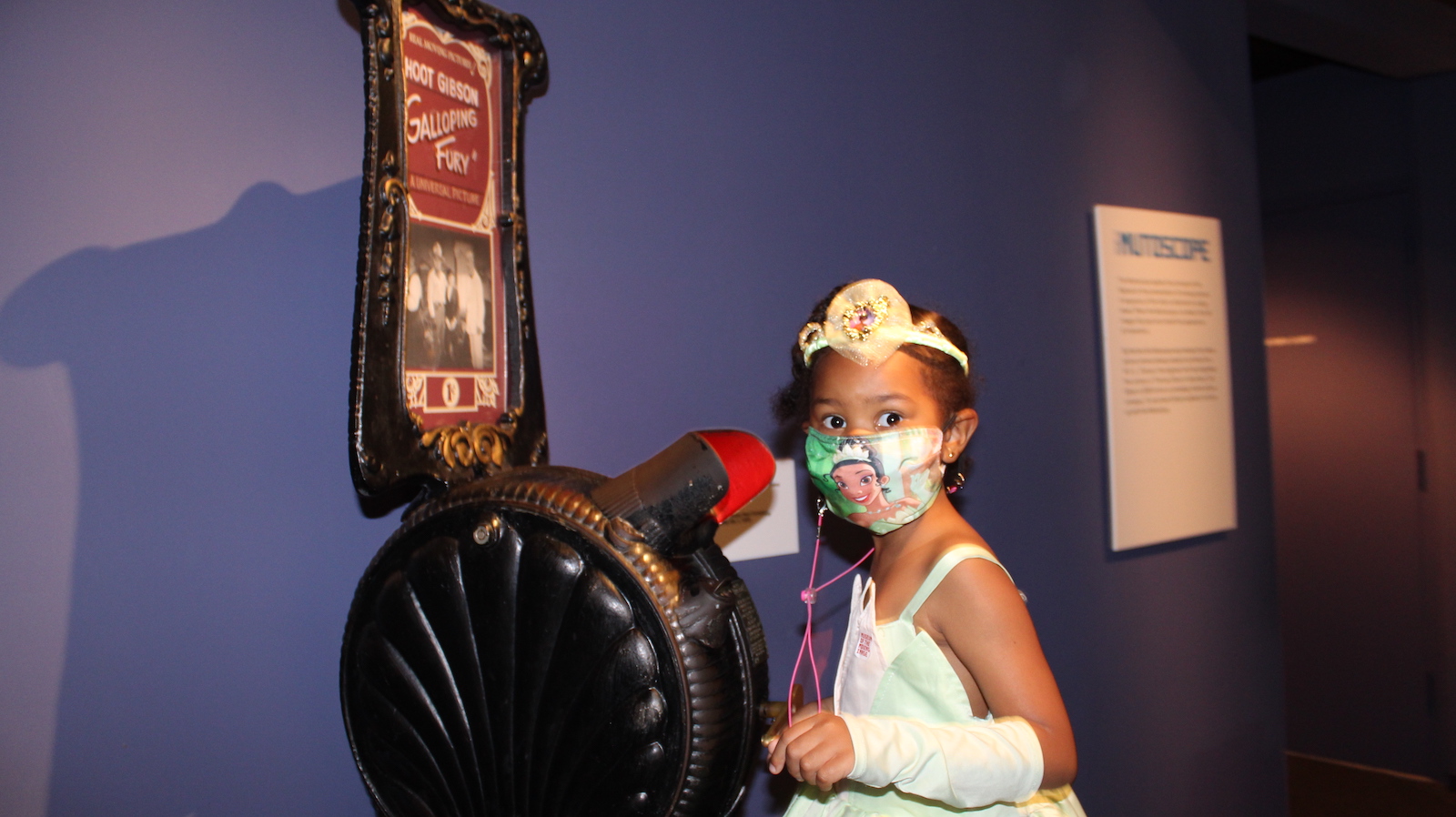 A little girl in a white dress and facemask looks at camera while standing next to a mutoscope device at Museum of the Moving Image