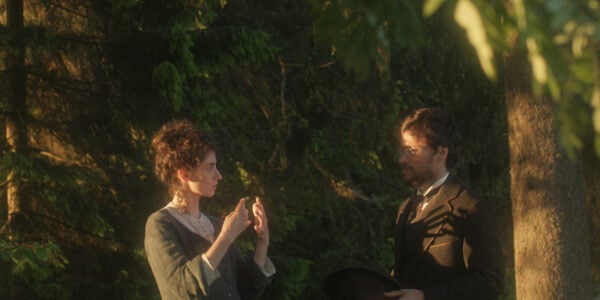 A woman and man in 19th century clothes talk to each other outside amidst a green forest setting