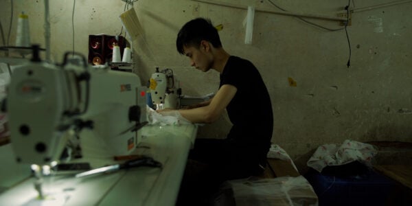 A young man sits over a sewing machine in a drab clothing factory